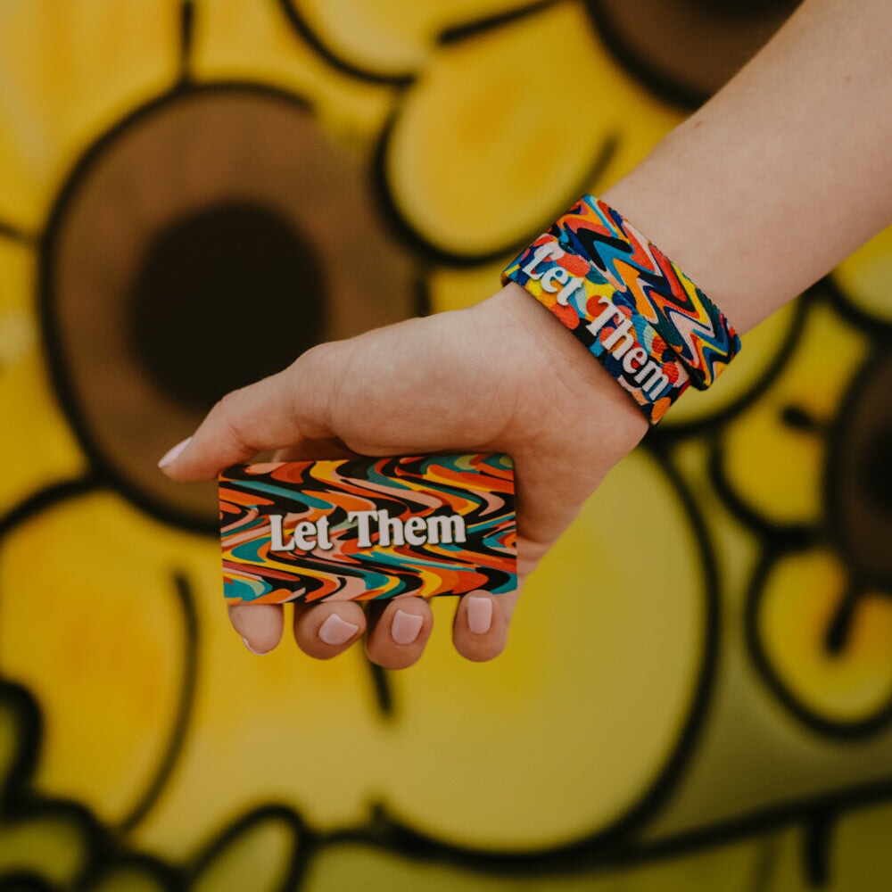 A hand with pastel nails holds a colorful "Let Them" card promoting self-acceptance, wearing the Let Them Bracelet. A yellow mural with large black circles appears in the background.
