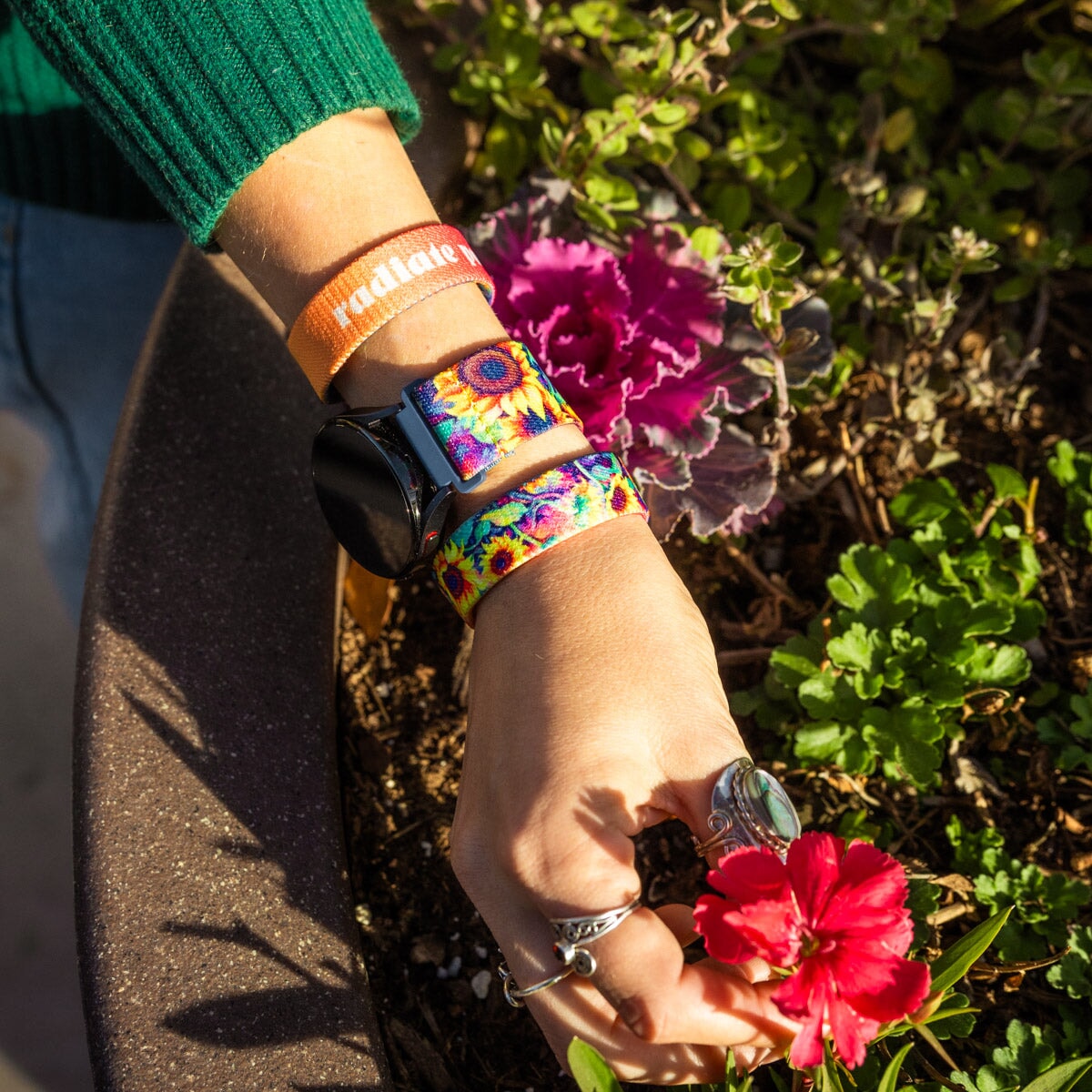 A close-up shot of a person's wrist wearing the Radiate Positivity watch band. The band features a vibrant sunflower design with bright yellow petals and dark centers. The person is gently touching a red flower in a garden, with green leaves and other plants visible in the background. The person also wears an orange bracelet with the text "radiate positivity" in white, and they have multiple silver rings on their fingers.