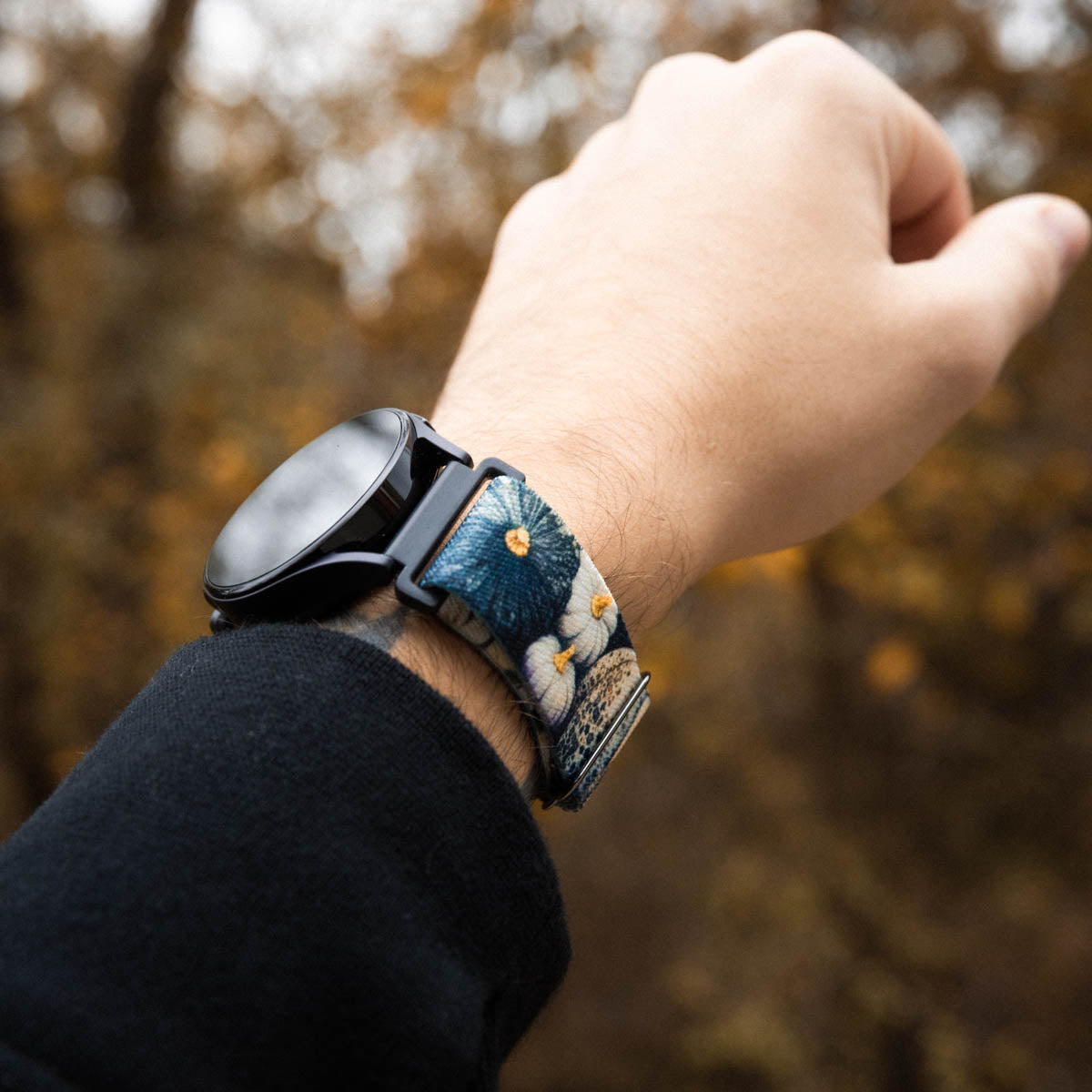 Lifestyle photo of Always Grateful Watch Band on a person's wrist featuring a floral pattern. The band displays detailed blue and white flowers with yellow centers, set against a textured background. The watch itself has a matte black finish, complementing the intricate design of the band. The scene is set outdoors with a blurred backdrop of autumnal trees, creating a warm, natural ambiance that highlights the watchband's artisanal look. The person's arm is dressed in a dark sleeve.