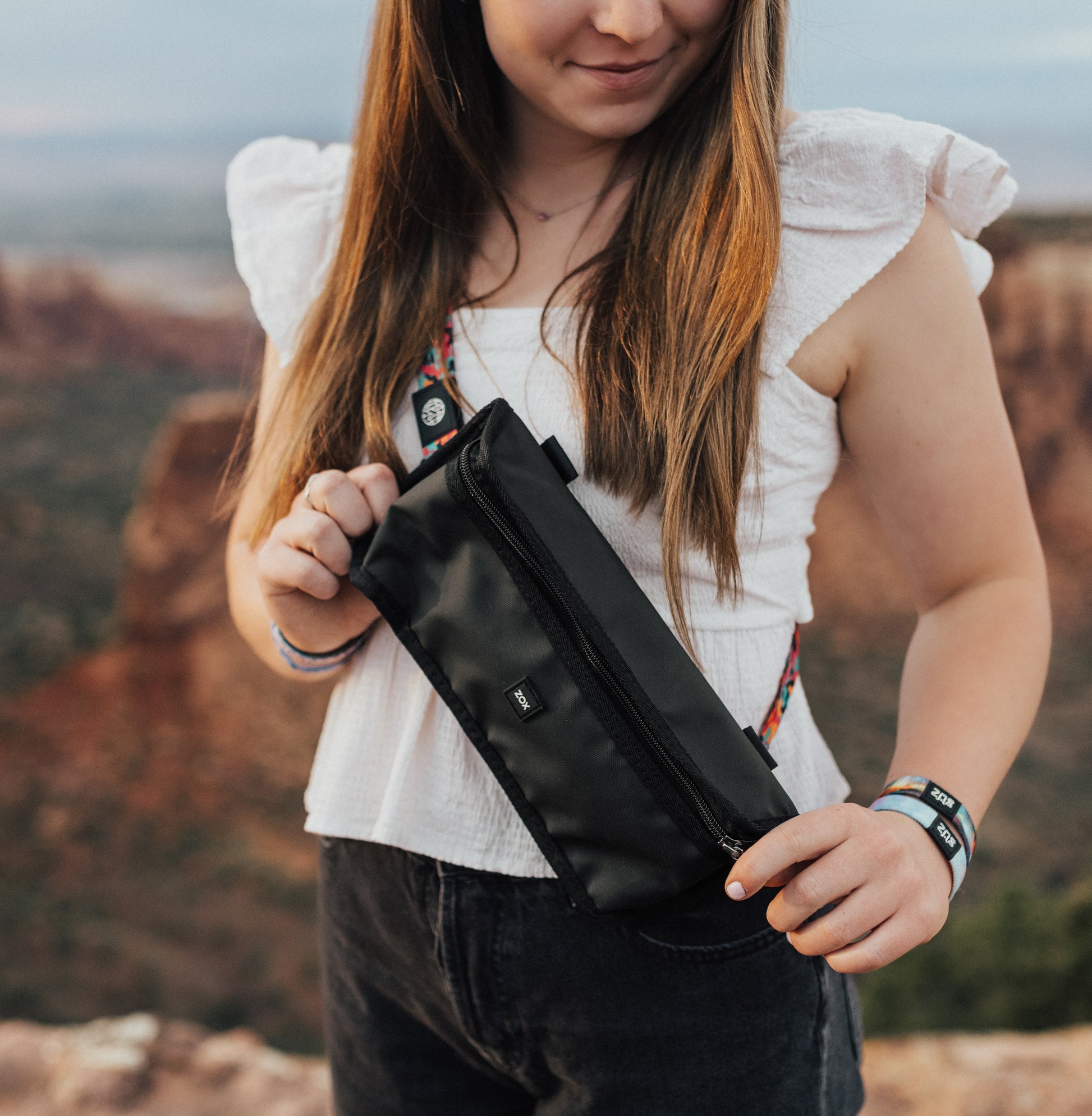 A person with long hair is wearing a black waist bag across their chest. The bag displays the "ZOX" logo on the front, and the colorful customizeable strap is visible. The person is outdoors, with a scenic background of a canyon or similar landscape.