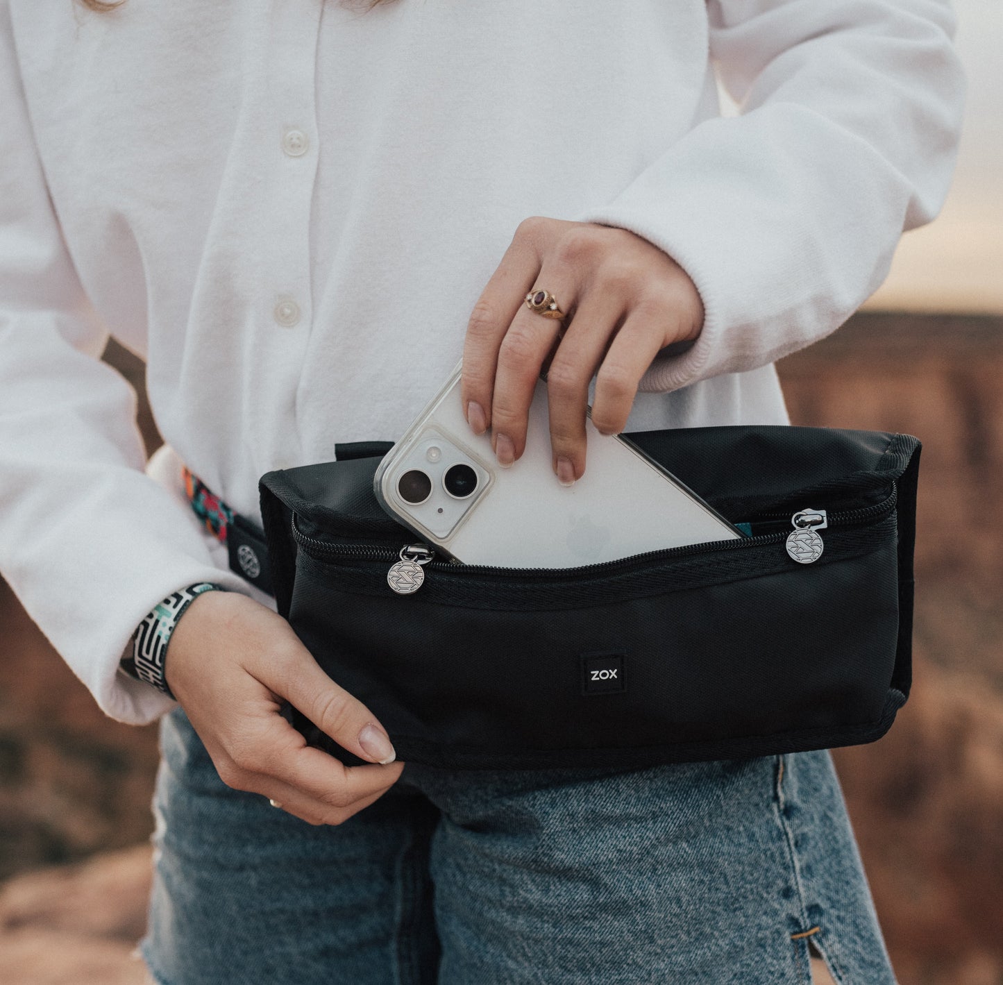 A person wearing a white shirt and jeans is holding a black waist bag with one hand while placing a smartphone into the zippered compartment with the other. The "ZOX" logo is visible on the front of the bag, and the person is standing outdoors with mountains behind them.