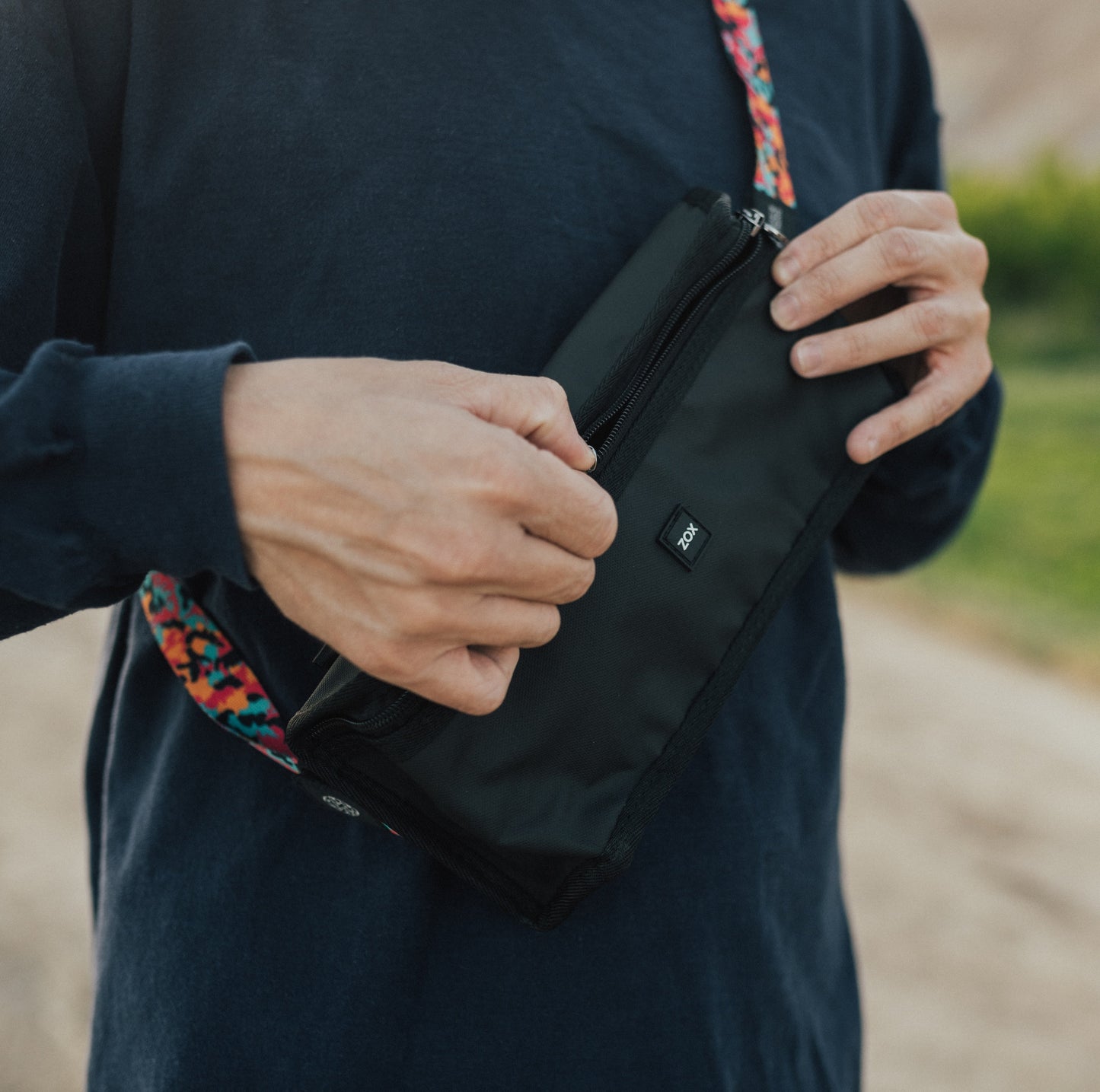 A person dressed in a dark long-sleeve shirt is adjusting a black waist bag worn across their chest. The bag has a visible "ZOX" logo on the front, and the strap features a colorful pattern. The background includes a hiking pathway surrounded by greenery.