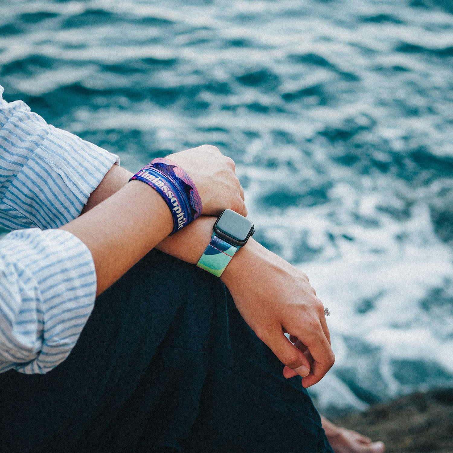 A person sitting by the ocean, resting their arms on their knees while wearing two bracelets. The Thalassophile bracelet on the left wrist features dolphins with a sunset ocean scene, and a Best is Yet to Come watchband is worn on the right wrist. The background is gentle ocean waves.