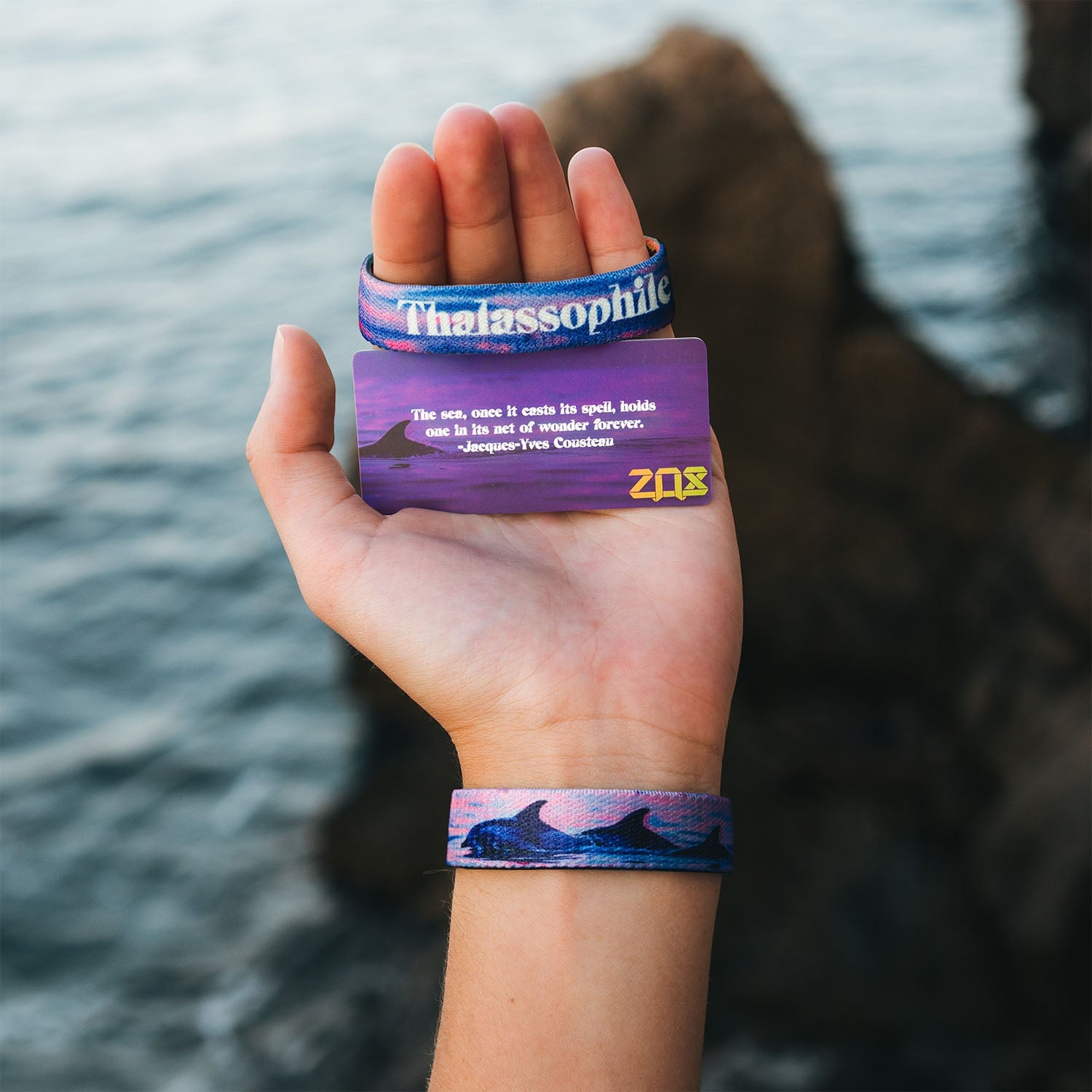 A hand holds the Thalassophile bracelet and its collector's card. The bracelet shows dolphins in a sunset scene, while the card displays a quote by Jacques-Yves Cousteau about the sea. The ocean and rocky shoreline are blurred in the background.