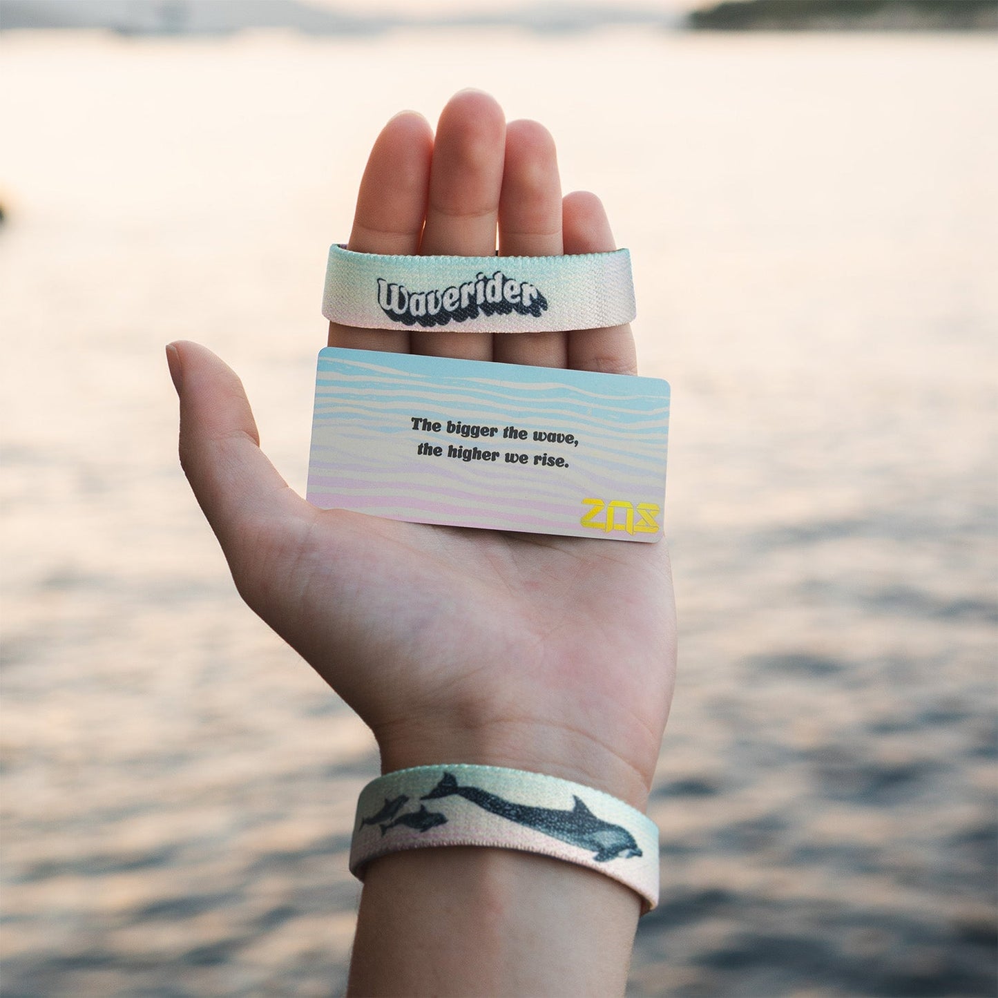 A hand is held up by the water, displaying the Waverider bracelet, which features dolphins on a pastel blue and pink background. The hand also holds the collector's card with the quote, "The bigger the wave, the higher we rise," and the gold "ZOX" logo in the lower corner. The calm water is visible in the background.