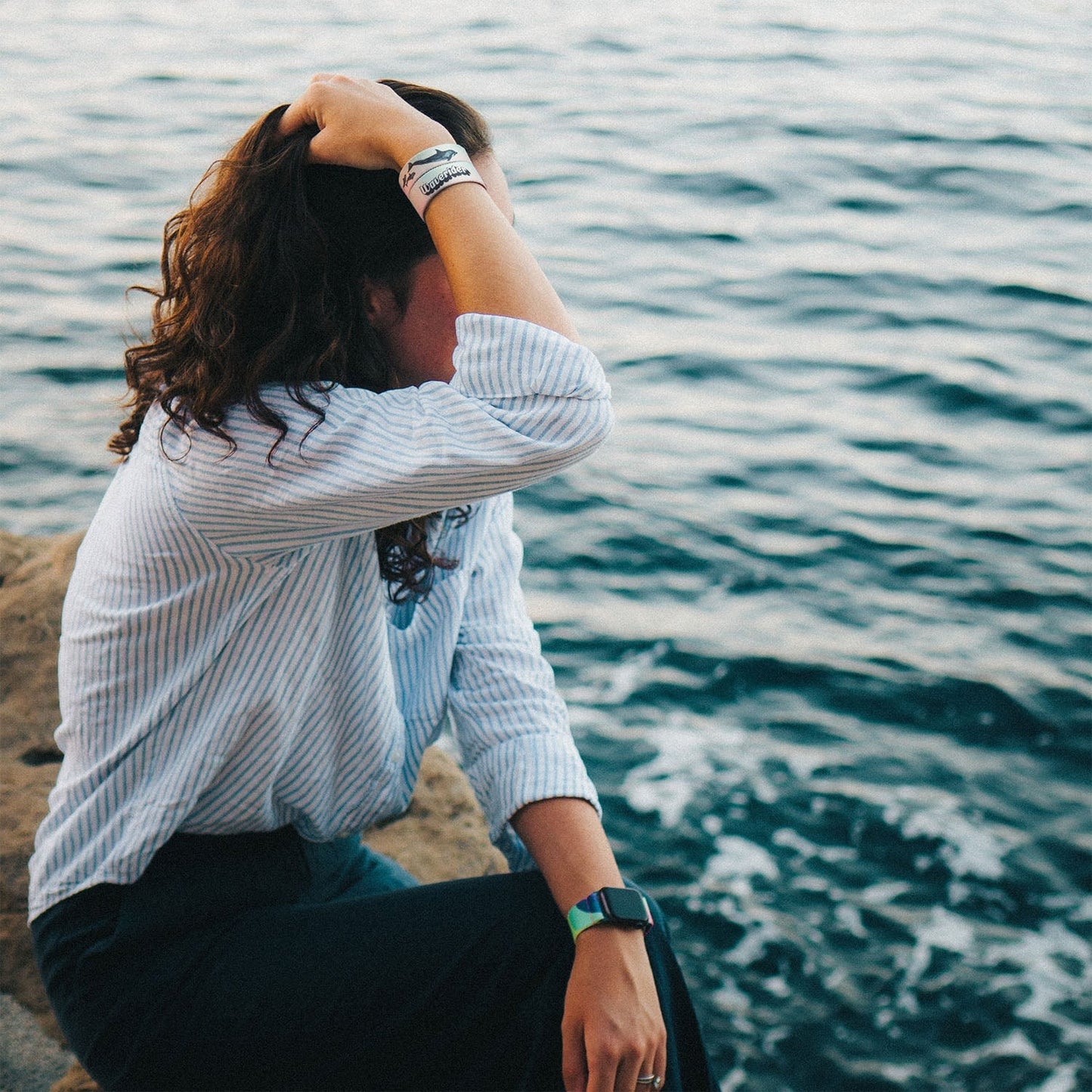 A person sits on a rocky shore, facing the ocean. They wear the Waverider bracelet on one wrist and a The Best is Yet to Come watchband on the other. The ocean waves in the background as the person runs their hand through their hair, wearing a striped shirt.