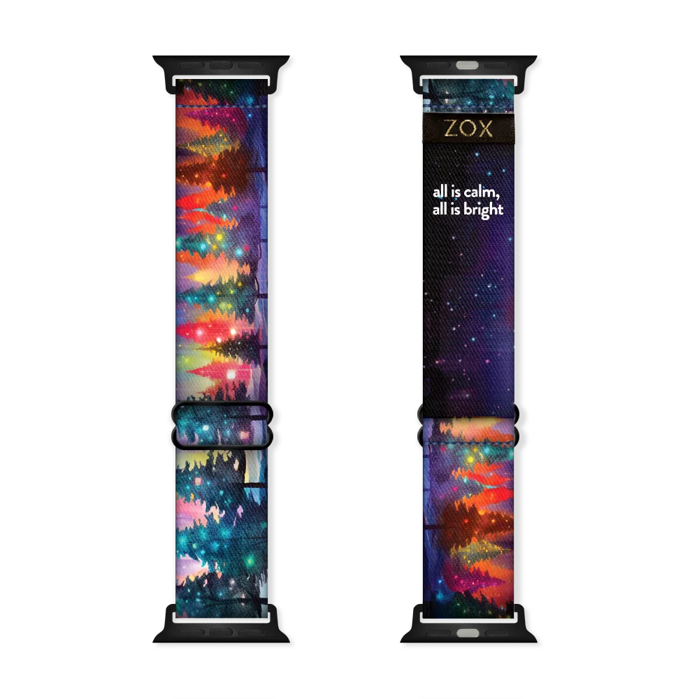 Apple Watchband render of the All is Calm, All is Bright design, showing both the outside (left) and inside (right) of the band. The outer side on the left features a colorful, night-time landscape with a forest of evergreen trees illuminated by glowing lights in hues of red, green, and yellow. The inside on the right is black with the phrase 'all is calm, all is bright' printed in white text. The ZOX logo is on a black tag sewn near the top.