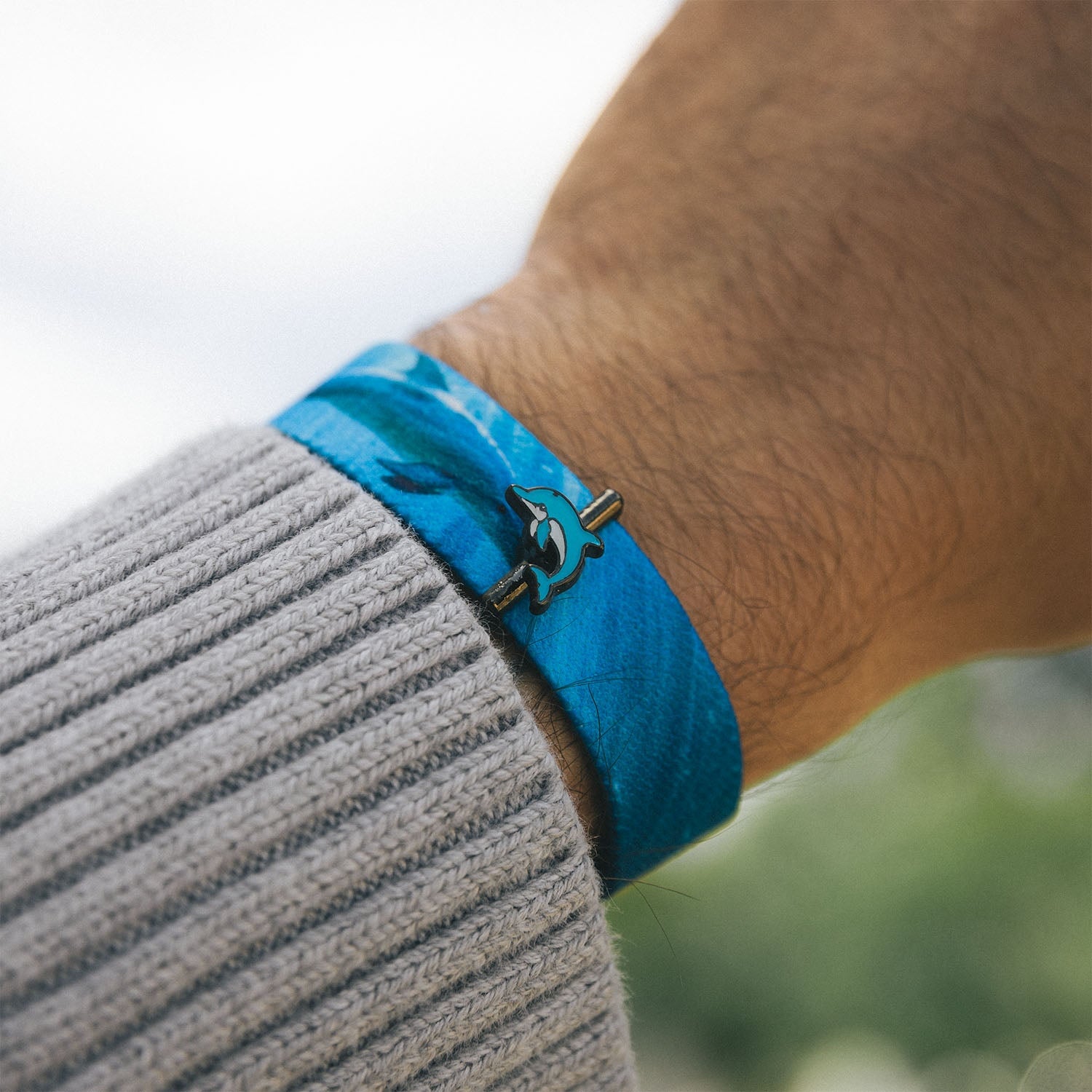 Lifestyle photo of Dolphin Charm which features a person's wrist wearing a blue fabric bracelet, with the same dolphin charm attached via the small metallic bar. The person is wearing a gray knit sweater, and the background is blurred, suggesting an outdoor environment.