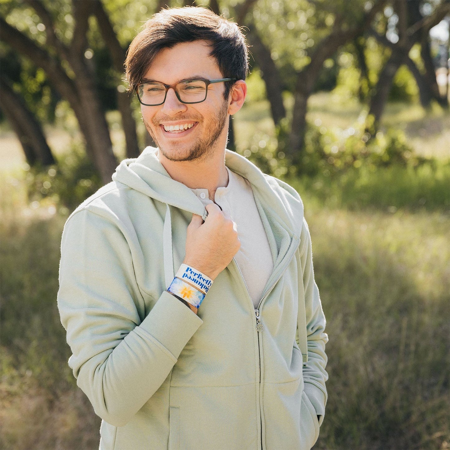 A man smiling outdoors while wearing a light green hoodie and glasses. He is standing in a sunlit field with trees in the background, showcasing the "Perfectly Balanced" bracelet on his wrist. The bracelet features a blue and orange mandala design with bold blue text.