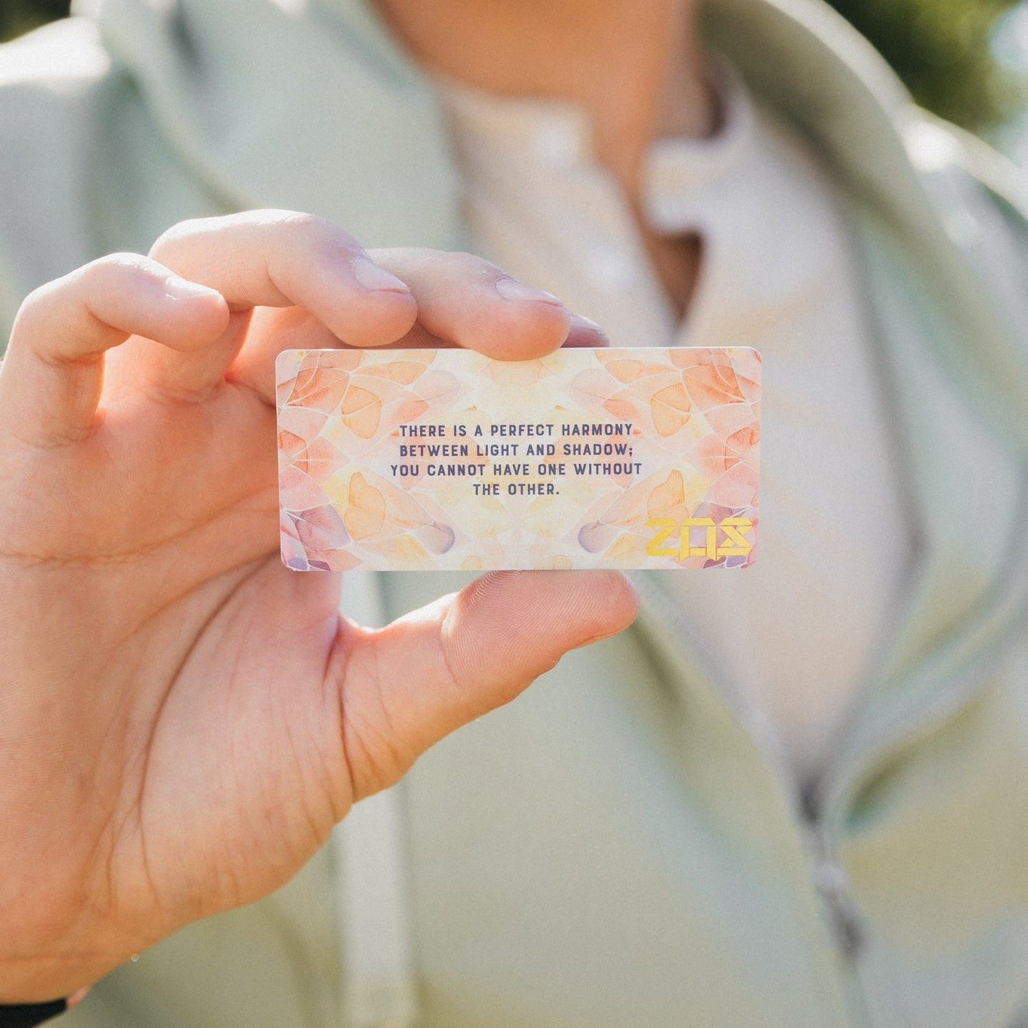 The man holds the collector’s card for the "Perfectly Balanced" bracelet. The card displays pastel watercolor petals in pink, orange, and beige. Blue text reads, "There is a perfect harmony between light and shadow; you cannot have one without the other," with a gold "ZOX" logo in the corner.