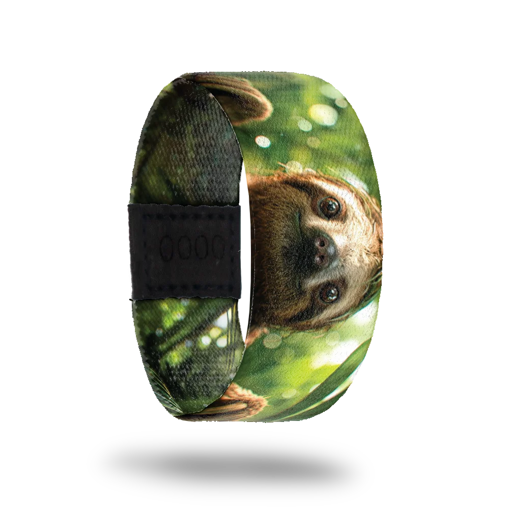 A bracelet featuring a close-up image of a smiling sloth's face set against a lush, green forest background with dappled sunlight filtering through the leaves. The sloth's expression is calm and content. A black rectangular patch with etched serial numbers is visible on one side of the strap.