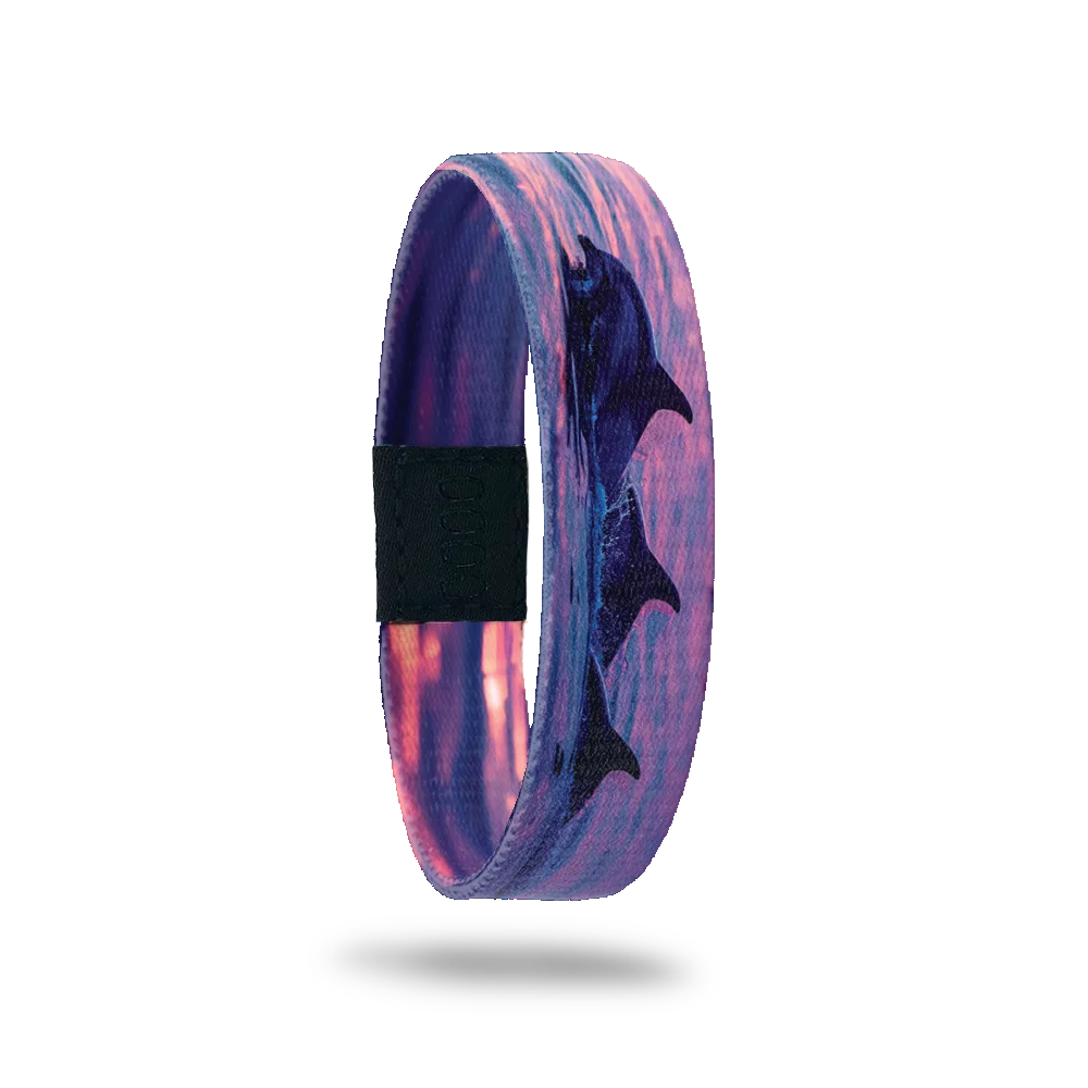 Product photo of the outside of the Thalassophile bracelet, featuring a vibrant sunset over the ocean with silhouettes of three dolphins leaping out of the water. The design captures shades of pink, purple, and orange, creating a serene and dynamic scene. A black rectangular patch with etched serial numbers is visible on one side of the single.