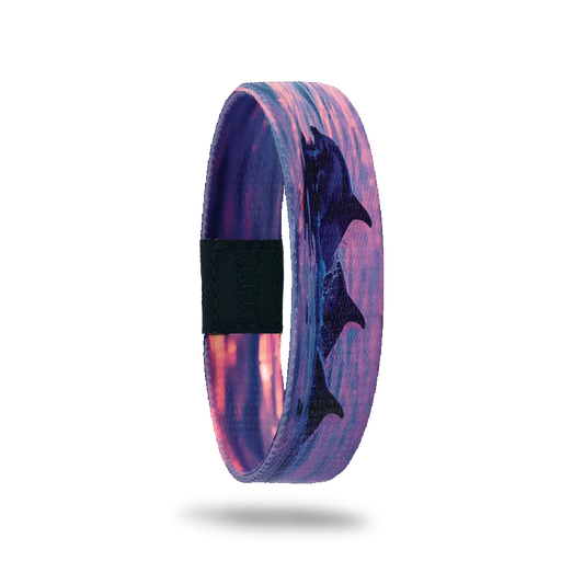 Product photo of the outside of the Thalassophile bracelet, featuring a vibrant sunset over the ocean with silhouettes of three dolphins leaping out of the water. The design captures shades of pink, purple, and orange, creating a serene and dynamic scene. A black rectangular patch with etched serial numbers is visible on one side of the single.