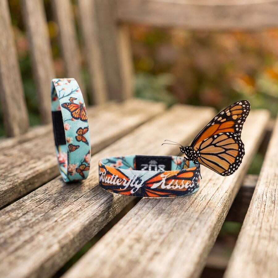 Butterfly-themed wristbands on a wooden bench with a butterfly perched on one of them, outdoors.