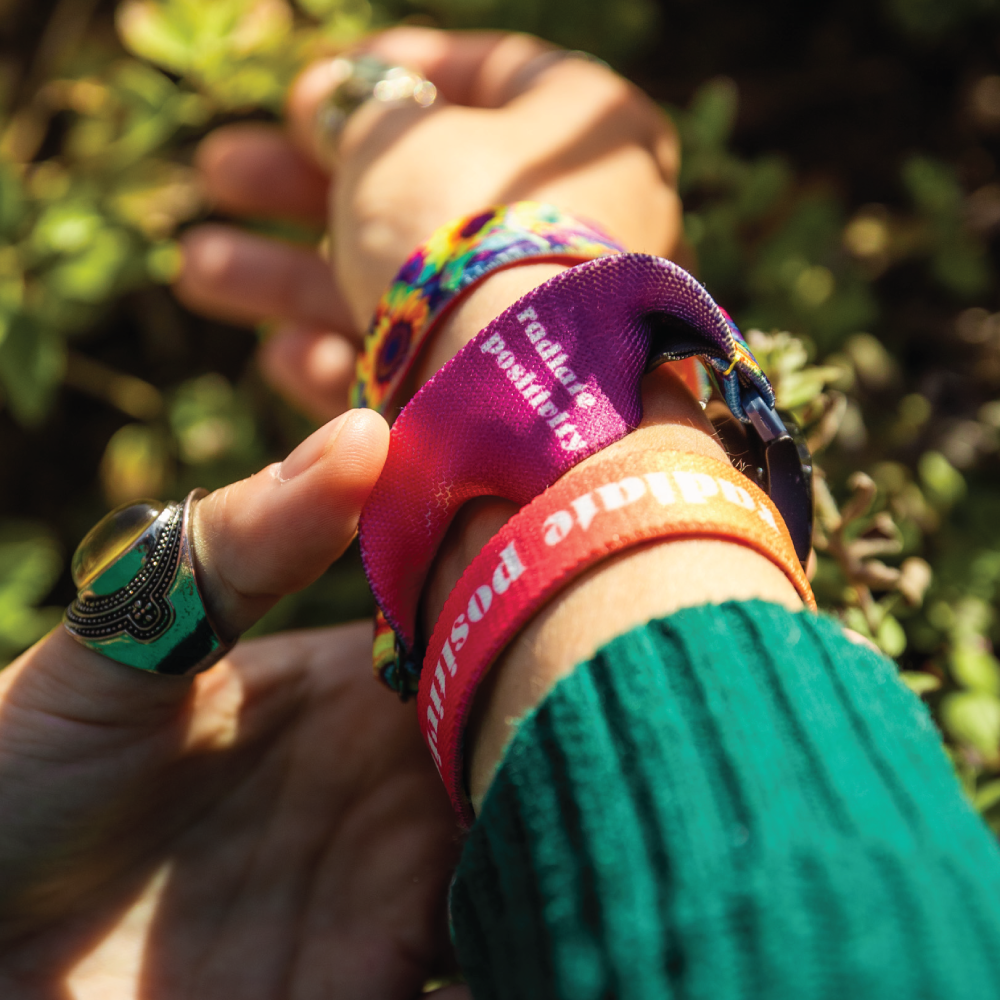 A close-up shot of a person's wrist showcasing the Radiate Positivity watch band. The band has a bright sunflower pattern on the outside, and the inside is visible, showing the gradient from purple to pink with the white text "radiate positivity." The person also wears an orange bracelet with the same text. They are gently holding their wrist, displaying rings on their fingers, and the background is filled with soft-focus greenery.