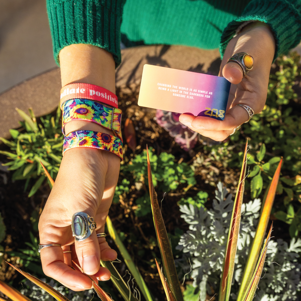 A person is shown holding the collector’s card for Radiate Positivity in one hand and wearing the matching watch band on their wrist. The card features a gradient from yellow to deep pink with white text that reads, "Changing the world is as simple as being a light in the darkness for someone else." The watch band displays a vivid sunflower design, and the person also wears an orange bracelet with the words "radiate positivity." The scene is set outdoors, surrounded by green plants and sunlight.