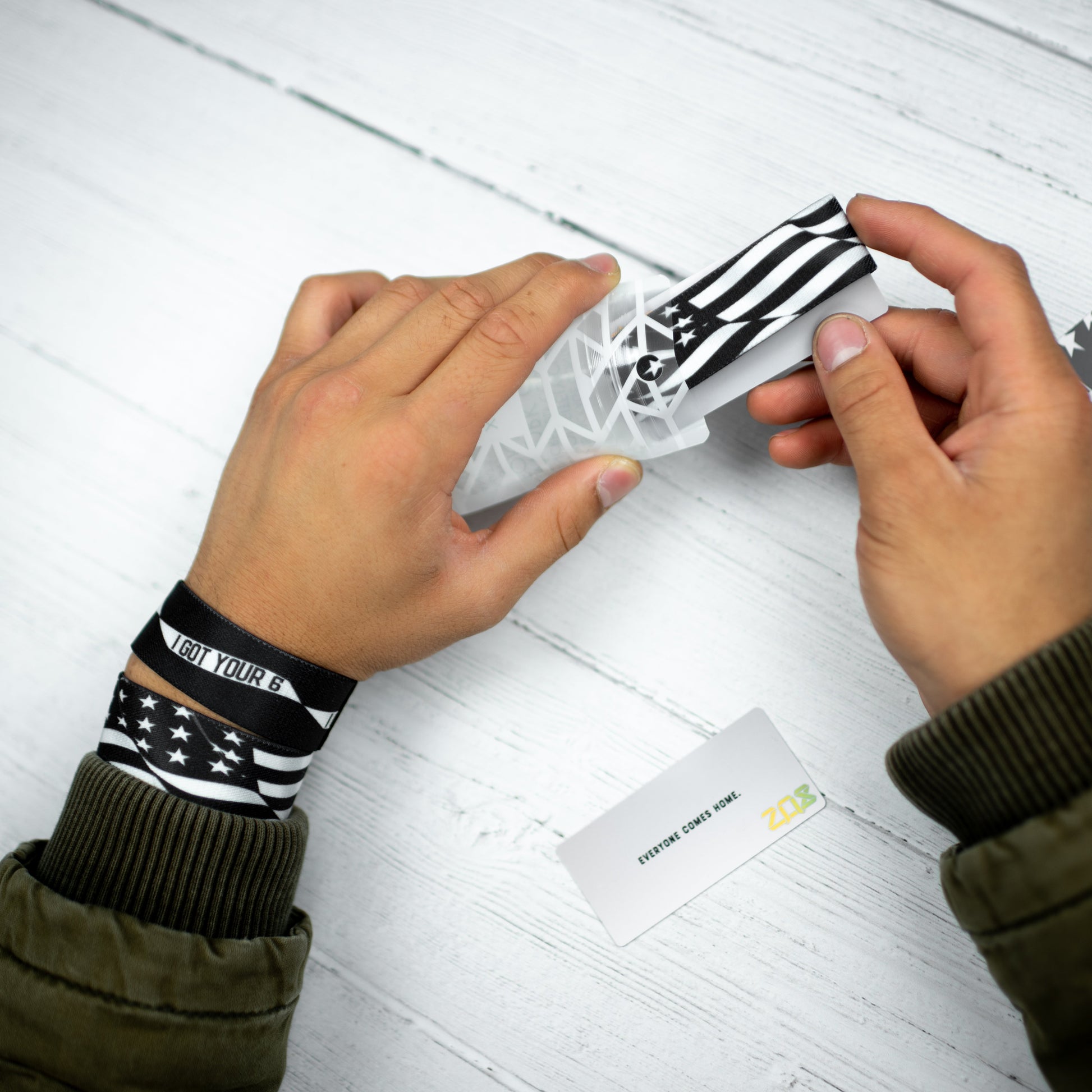 Studio Image of hands holding card with black and white United States flag on the card with 2 I Got Your Six straps on their wrist